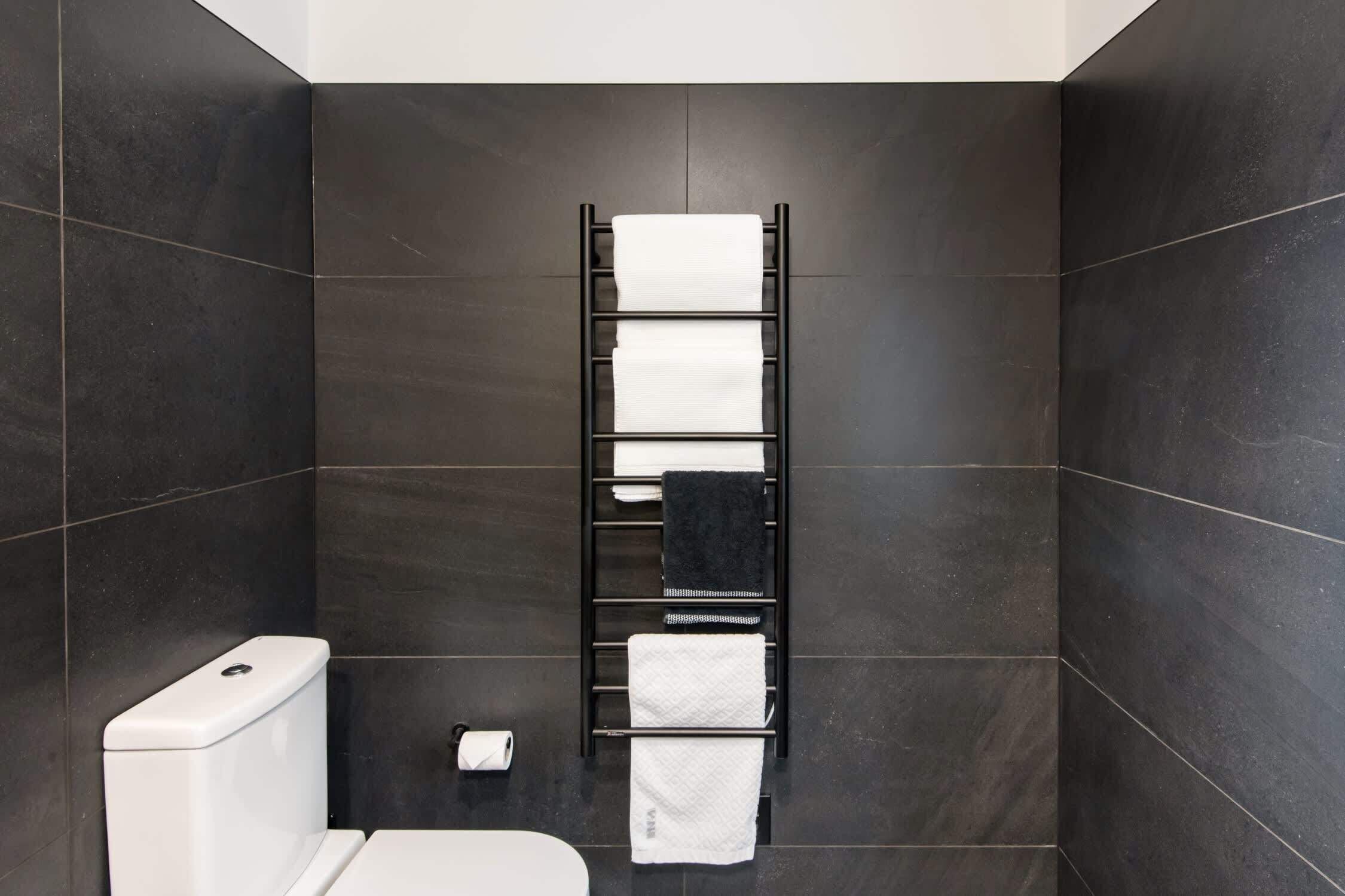 A bathroom with a toilet, a towel rack with multiple towels, and a toilet paper dispenser.
