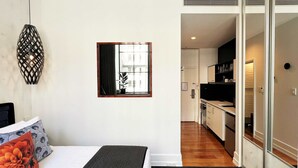 A bedroom with a bed, a nightstand, a wall-mounted lamp, and a view into the kitchen. - Stylish Studio with Full Kitchen on Hobson Street (Auckland Central)