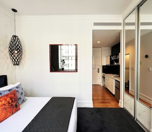 A bedroom with a bed, a nightstand, a wall-mounted lamp, and a view into the kitchen. - Stylish Studio with Full Kitchen on Hobson Street (Auckland Central)