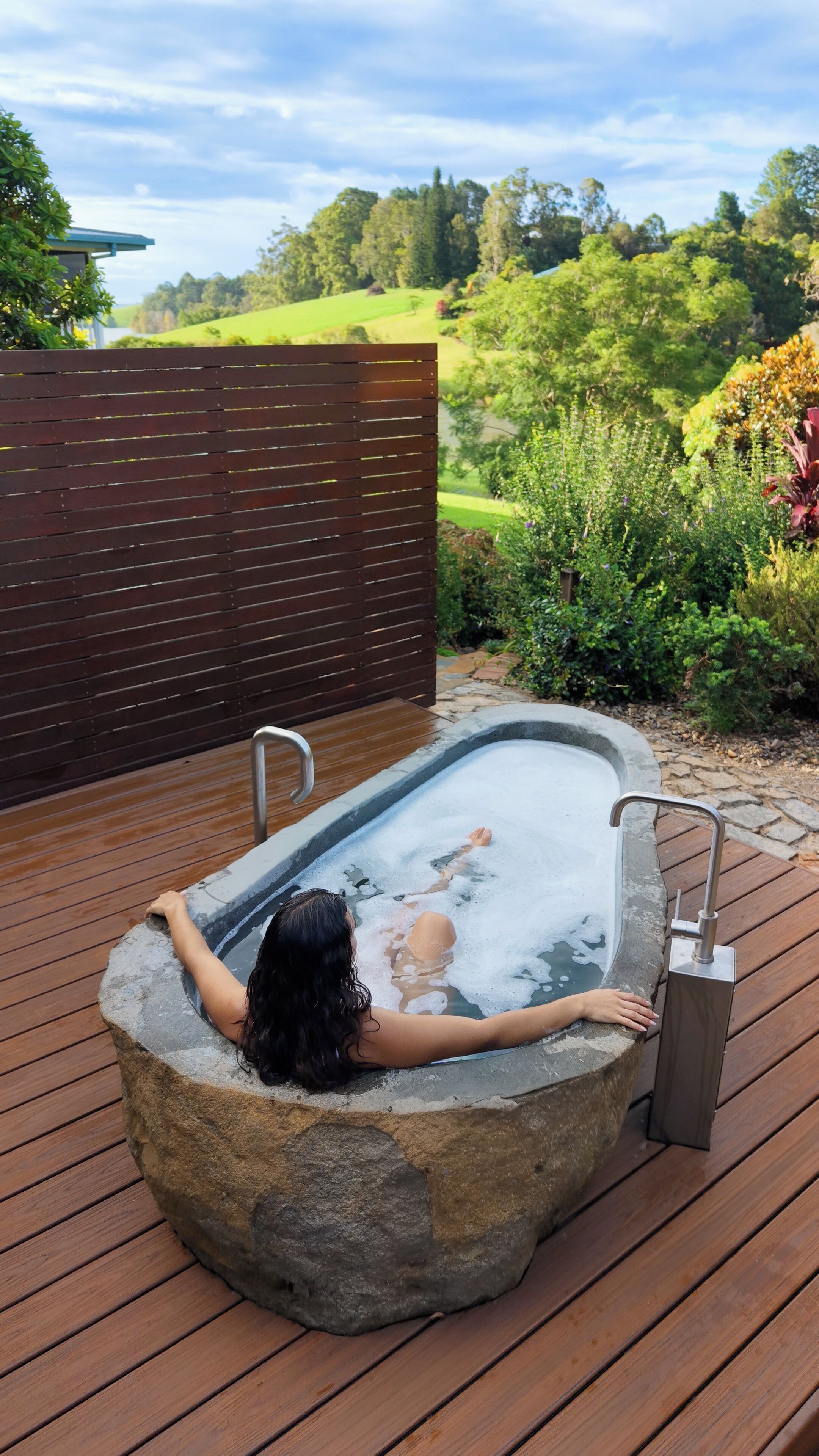 A person relaxing in a stone hot tub on a wooden deck with a garden view.