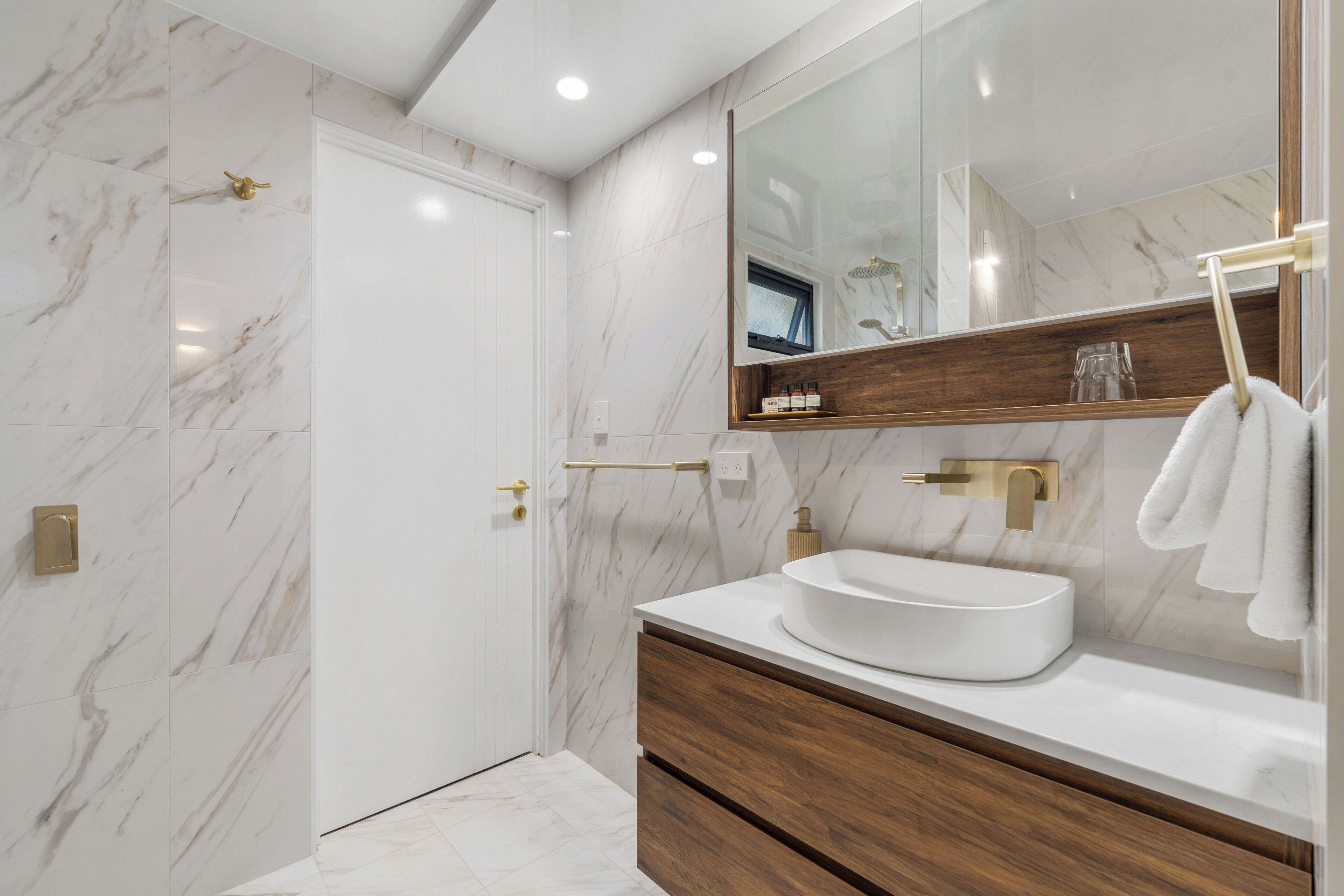Une salle de bain moderne avec un mur en marbre blanc, un meuble-lavabo en bois, un grand miroir et un lavabo blanc.