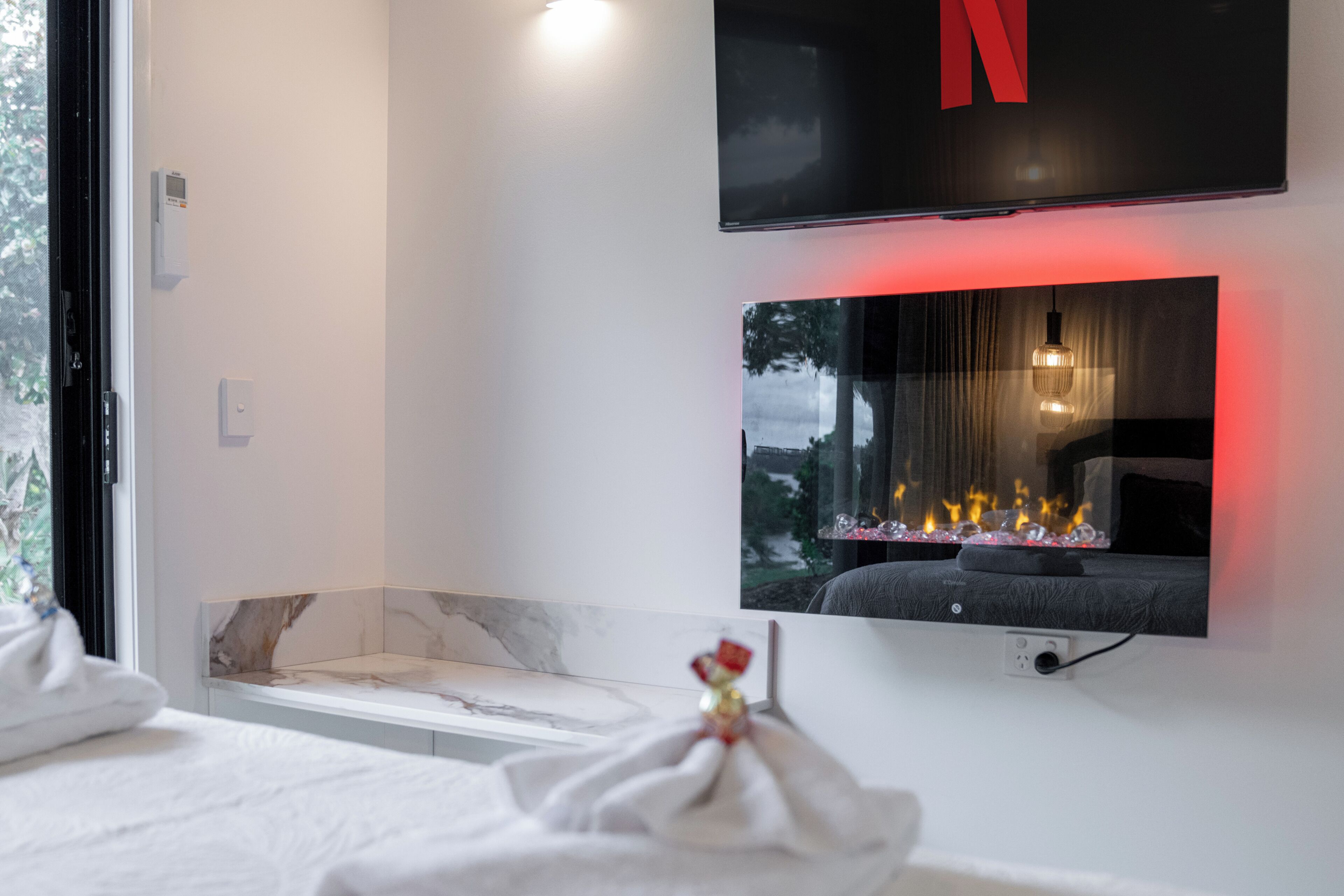 A modern bathroom with a large mirror, a flat-screen TV mounted above it, and a fireplace below.