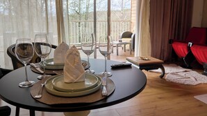A dining table set with plates, glasses, and cutlery, a remote control, and a view of the outdoors through large windows. - Luxury loveroom with private Jacuzzi, sauna, cinema corner & privacy area (Saint-Privat)