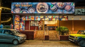 A restaurant with a signboard displaying various dishes, a staircase leading to the entrance, and two parked vehicles. - Karar Gardens (Kumily)