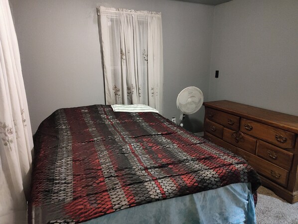 A bedroom with a bed, a dresser, a fan, and a window with curtains.