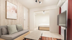 Uma sala de estar moderna com um sofá, uma mesinha, uma televisão e uma cama ao fundo.