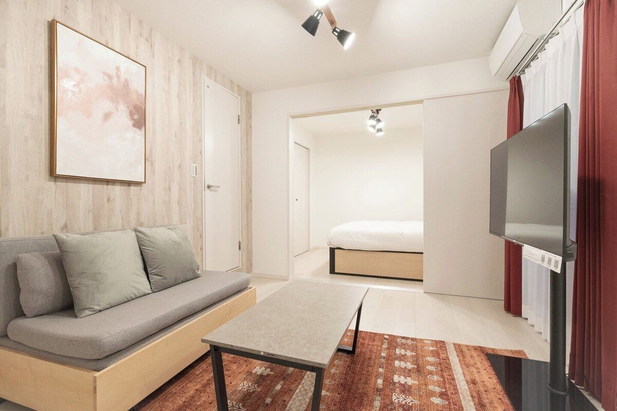 Uma sala de estar moderna com um sofá, uma mesinha, uma televisão e uma cama ao fundo.