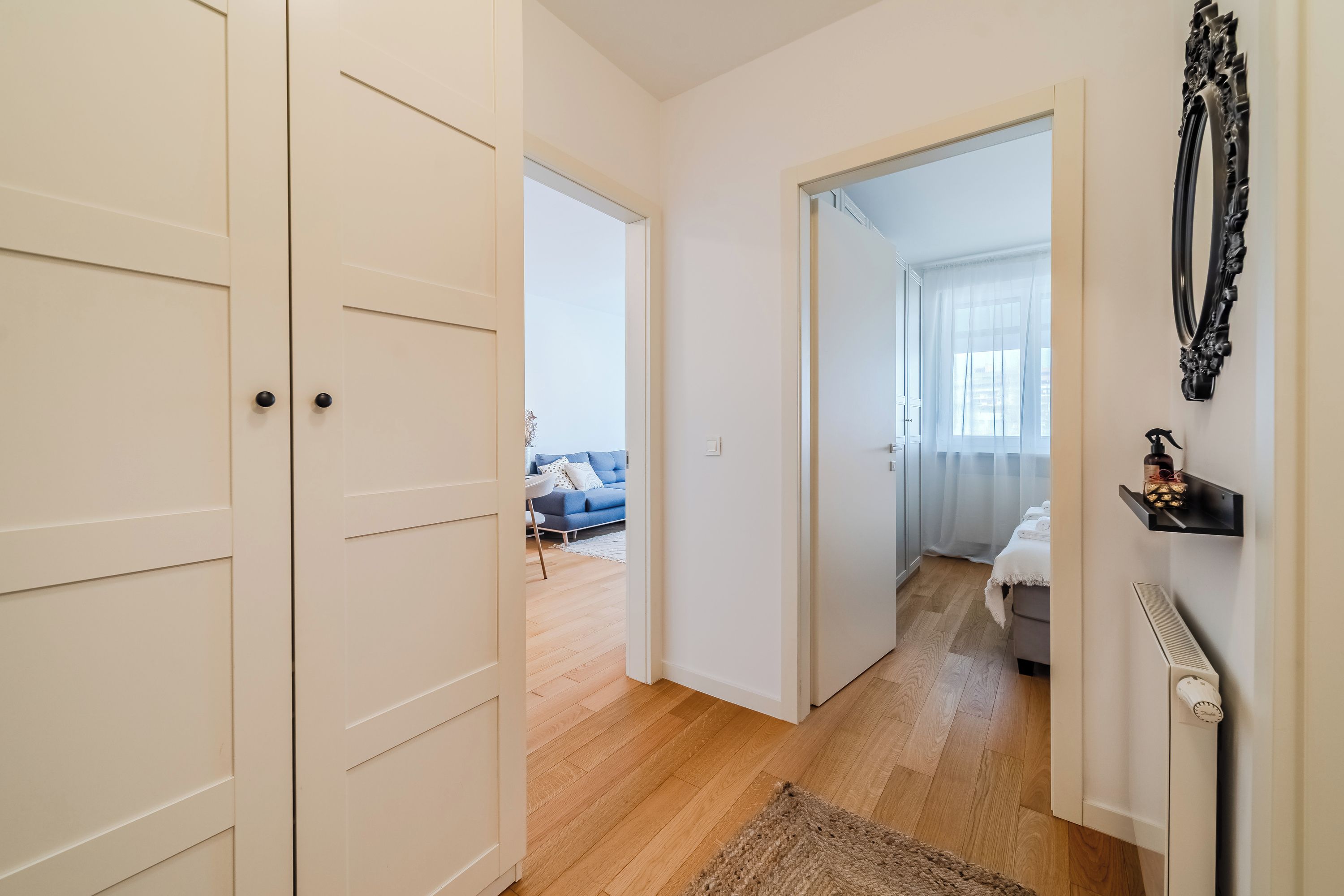 Un corridoio con pavimento in legno, pareti bianche e una porta che conduce a una camera da letto con un letto e una finestra.