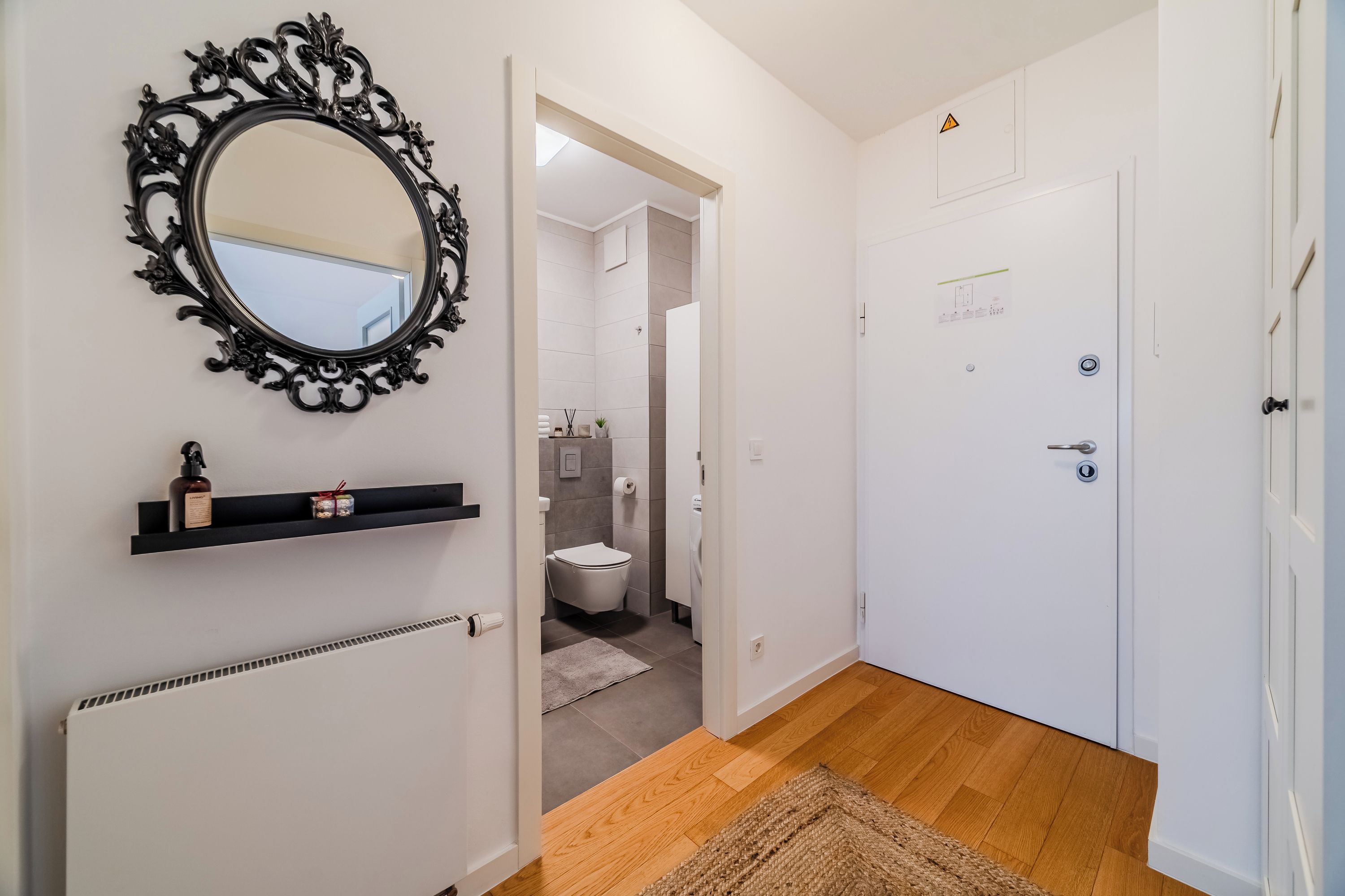 Een hal met een spiegel, een radiator, een plankje met toiletartikelen, een deur en een badkamer die door een open deur zichtbaar is.