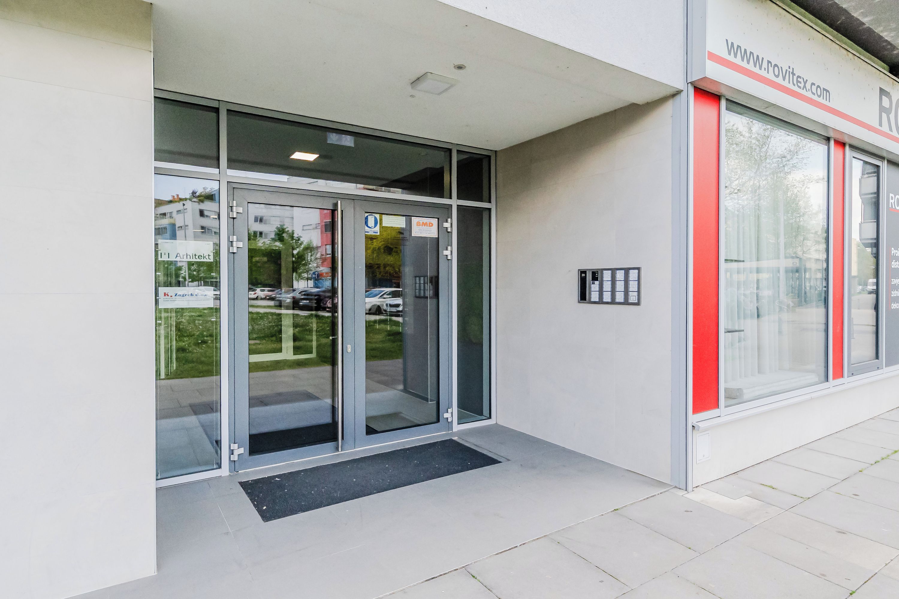 Ingresso di un edificio con porte in vetro, un indirizzo web e un cartello con indicazioni per il parcheggio.
