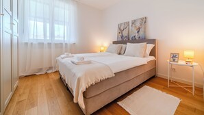 Un dormitorio con cama, mesitas de noche, cortinas translúcidas en la ventana y una obra de arte en la pared.