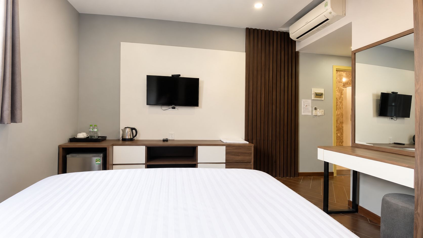 A hotel room with a bed, a desk with a flat-screen TV, a minibar, and a large mirror.