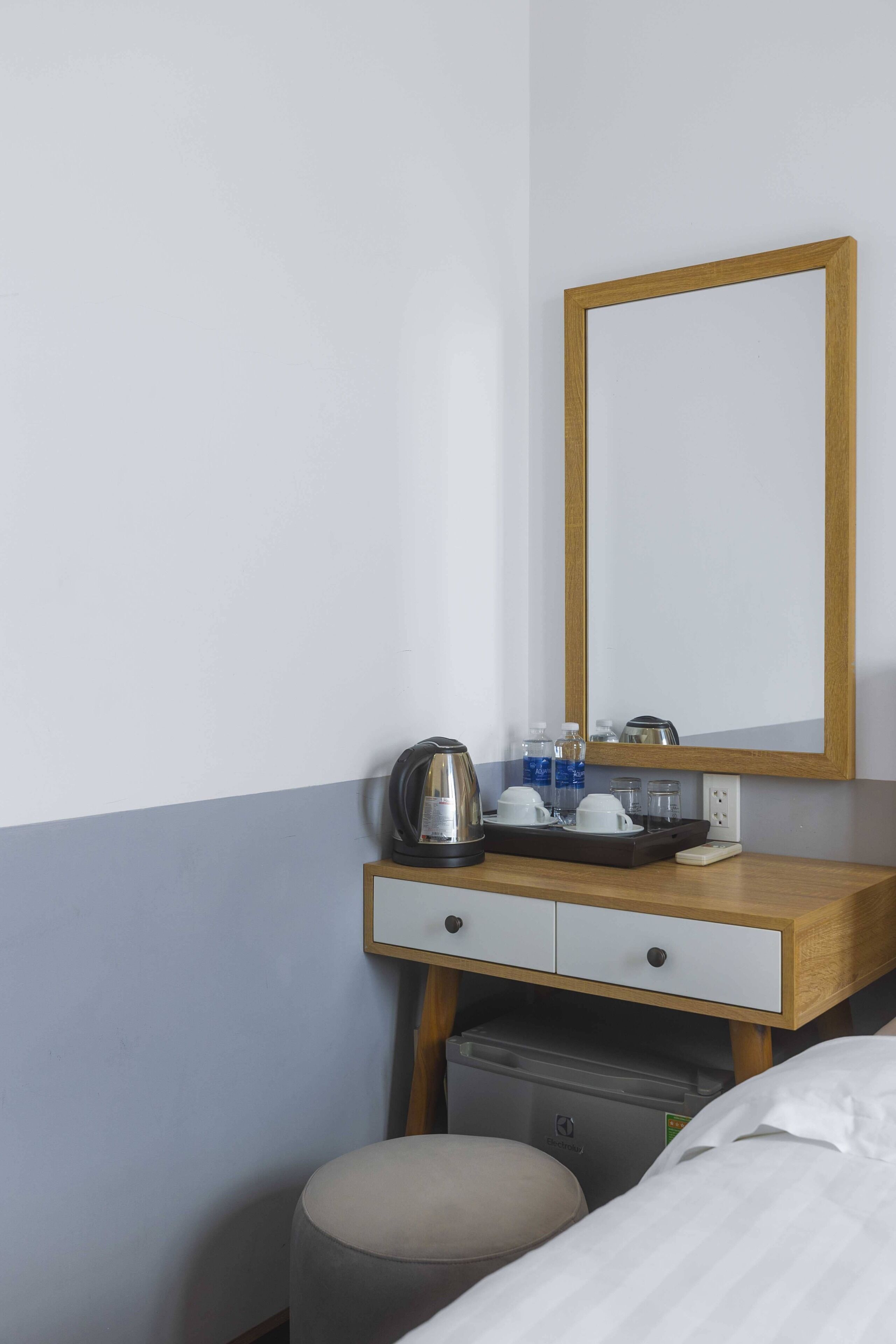 Habitación de hotel con un escritorio de madera, un espejo, una tetera, tazas y un cubo de basura pequeño.