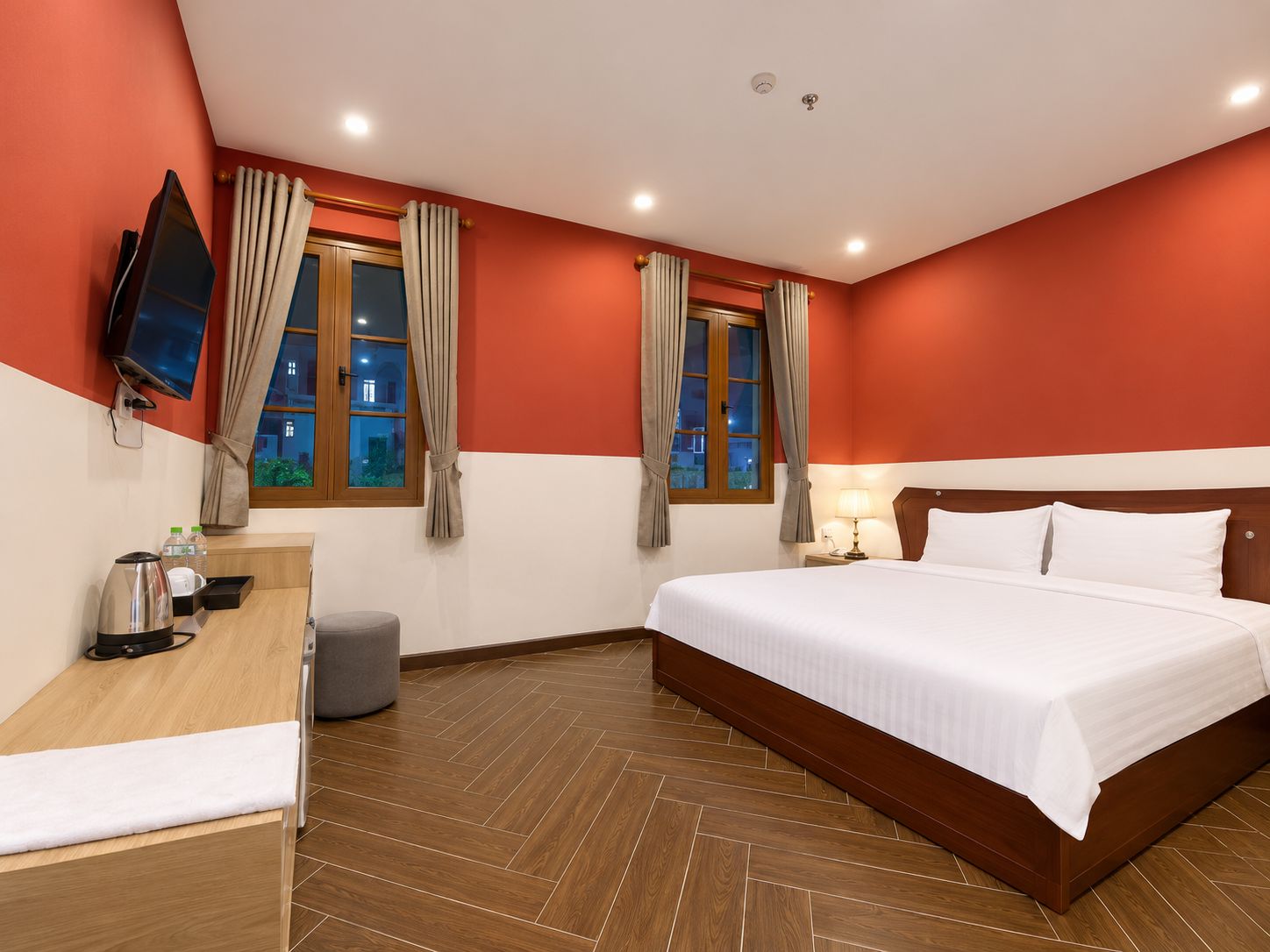 Habitación de hotel con cama, escritorio con hervidor, televisor y dos ventanas con cortinas.
