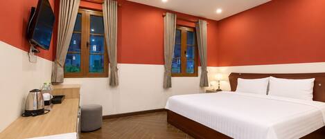 Habitación de hotel con cama, escritorio con hervidor, televisor y dos ventanas con cortinas.