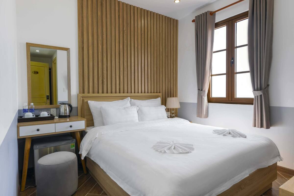 A hotel room with a large bed, a wooden headboard, a bedside table with a lamp, a mirror, and a window with curtains.