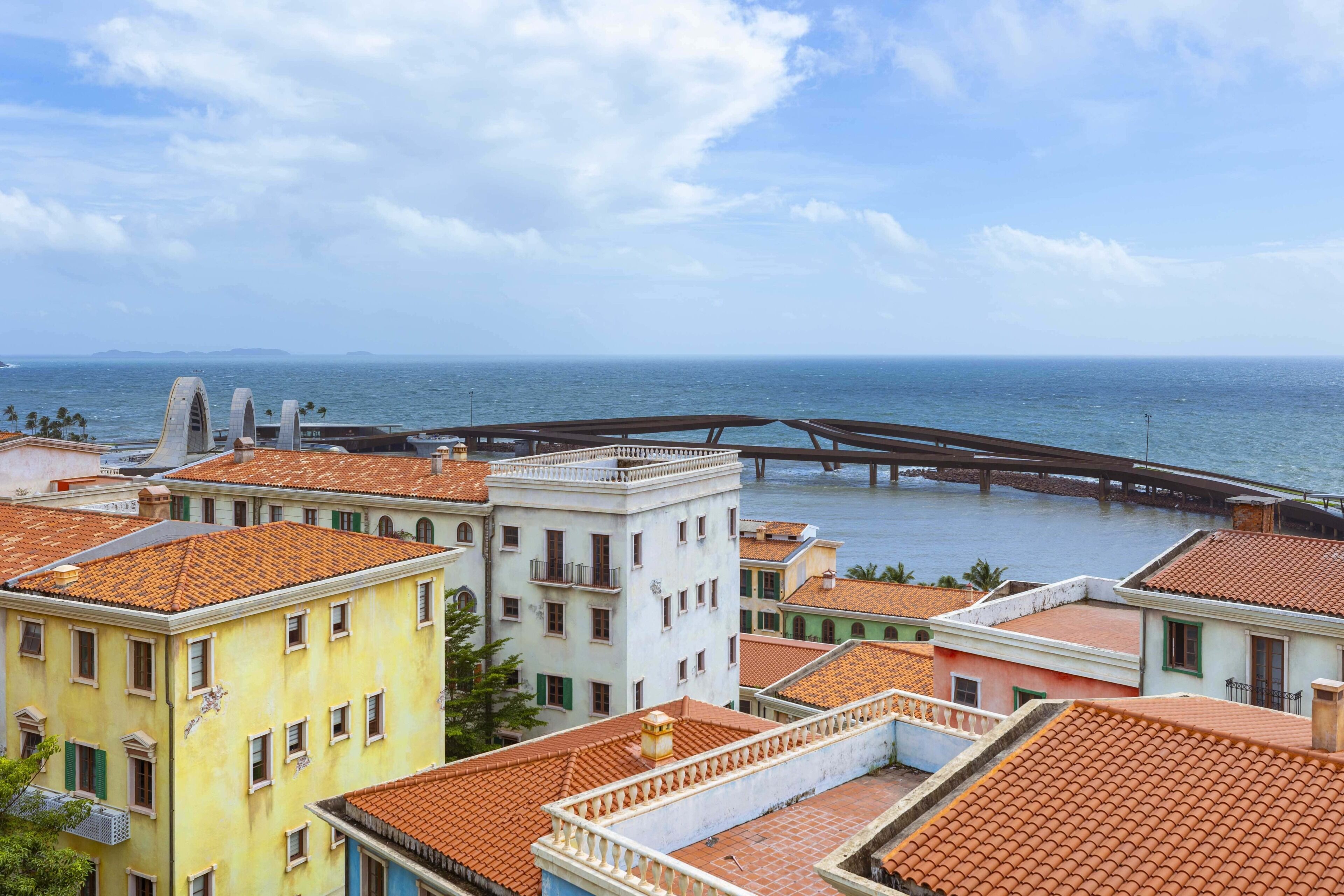 A coastal cityscape with terracotta-roofed buildings, a bridge over water, and a clear sky.