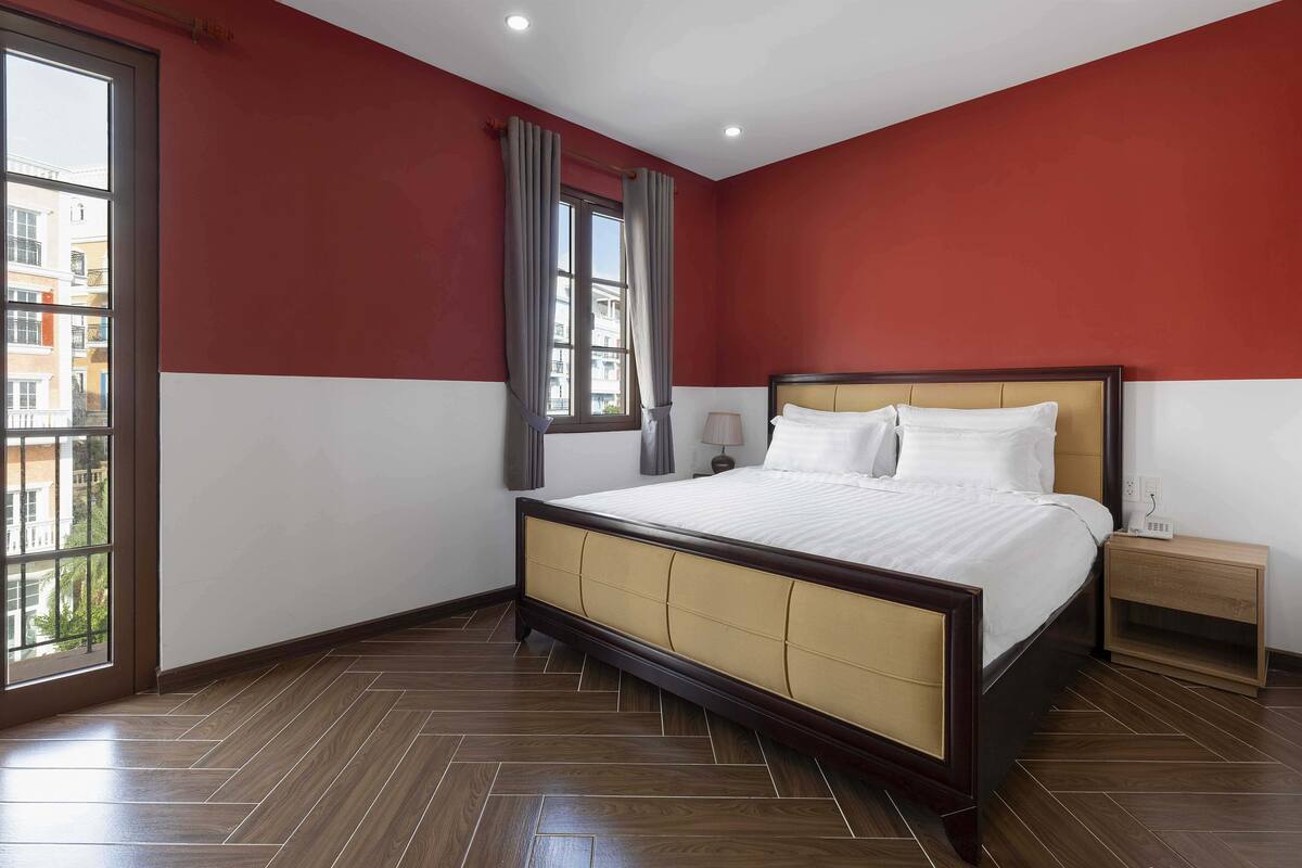 A hotel room with a large bed, a nightstand, a window with curtains, and a view of buildings outside.