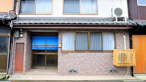 A two-story residential building with a tiled roof, a blue awning over the entrance, and an air conditioning unit on the second floor. - KAKENKAN (Miyazu)