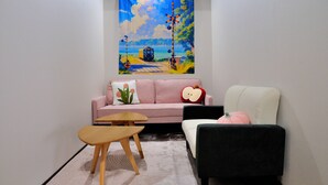 A living room with a pink sofa, a black armchair, a small wooden table, and a tapestry depicting a train on tracks. - KAKENKAN (Miyazu)