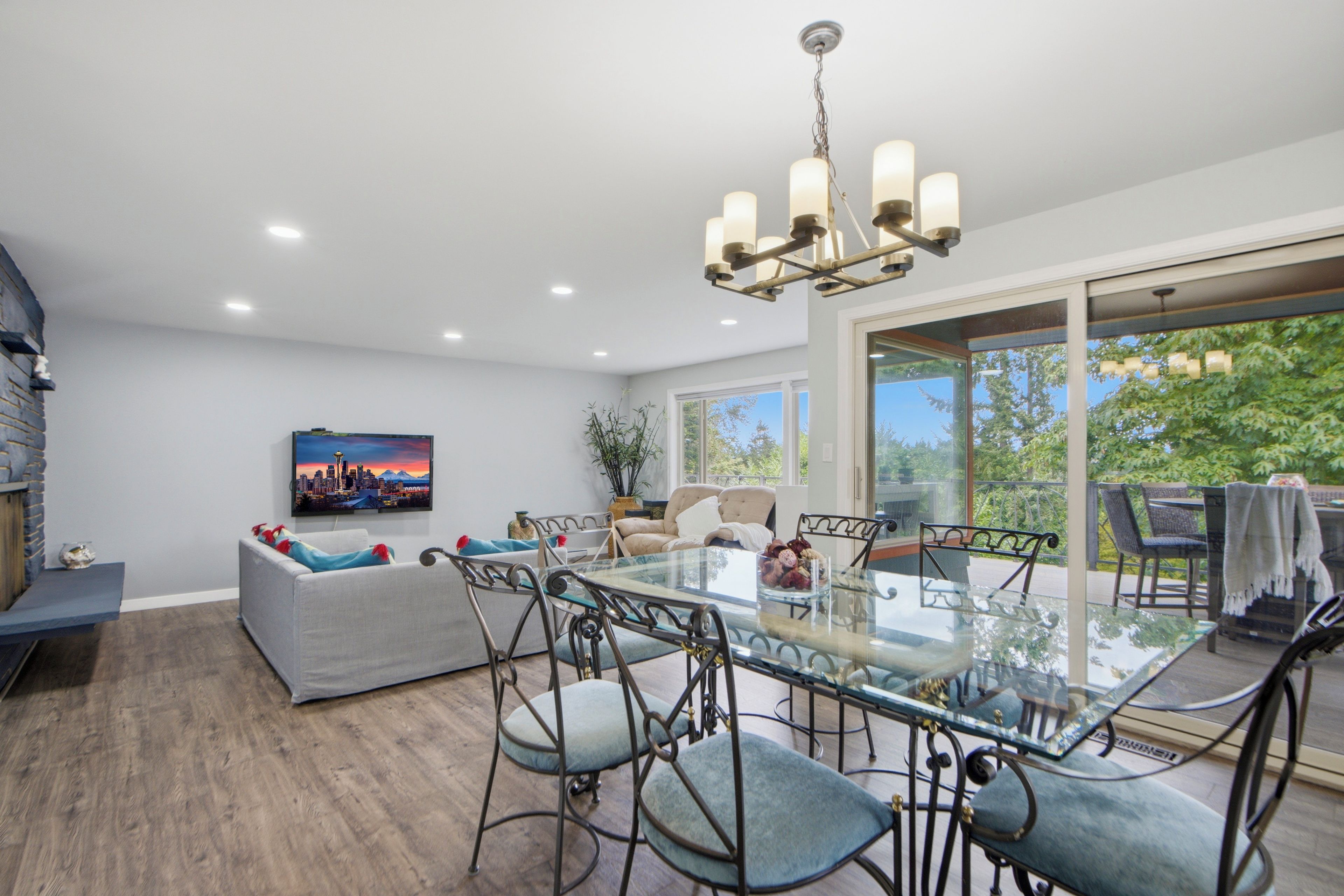 A spacious living area with a glass dining table surrounded by chairs, a sofa with decorative pillows, a television mounted on the wall, and a large window offering a view of the outdoors.