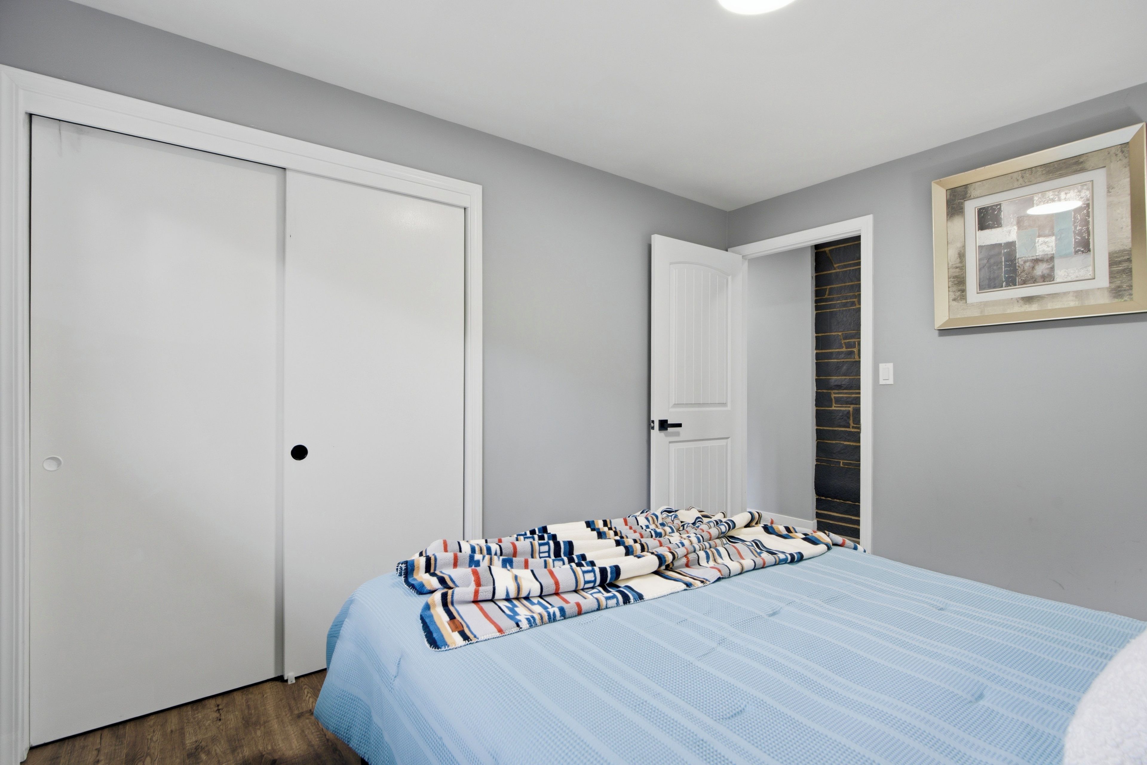 A bedroom with a bed, two doors, a framed picture on the wall, and a wooden floor.