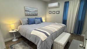 Une chambre à coucher comprenant un lit, des tables de chevet, un banc, un appareil de climatisation et une fenêtre avec des rideaux.