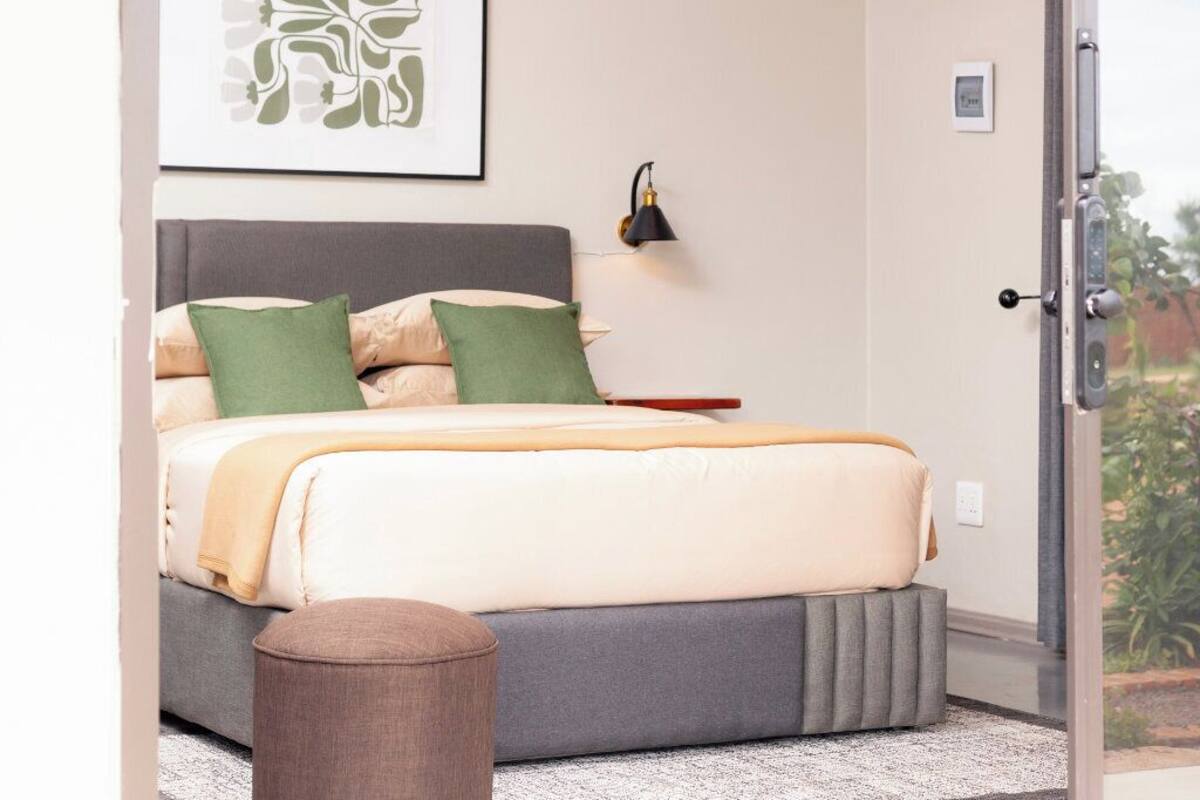 Un dormitorio con cama, almohadas, una lámpara de noche, una puerta y vistas al verde exterior.