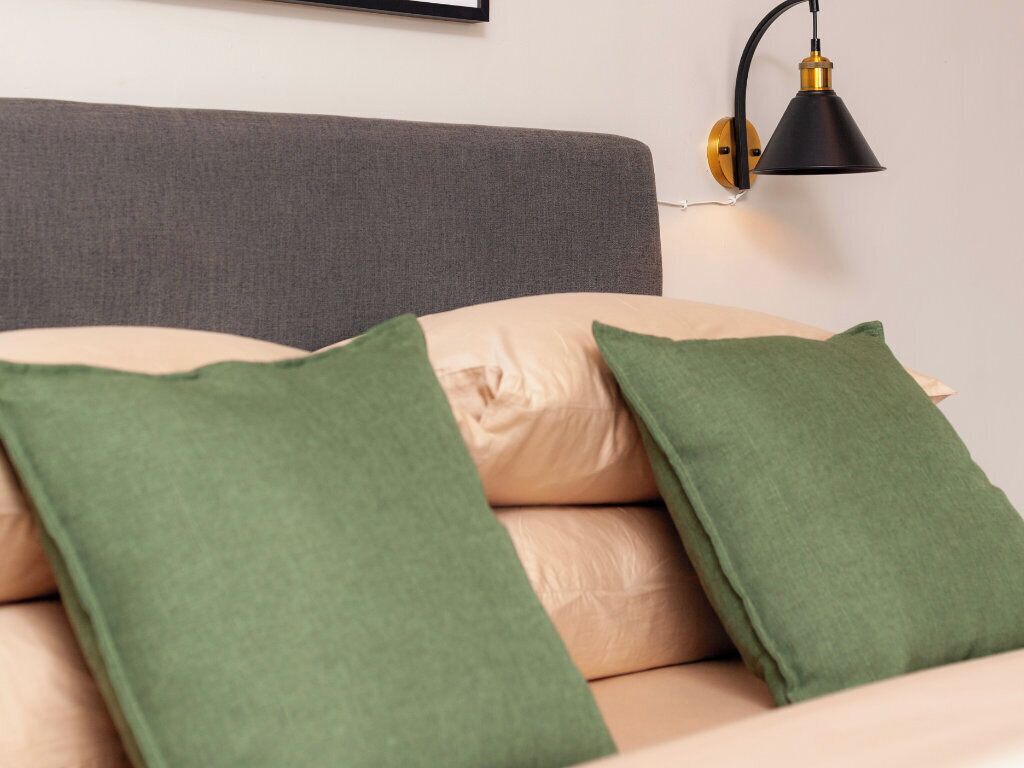 Een bed met groene en beige kussens, een grijze hoofdbord en een zwart wandlamp.