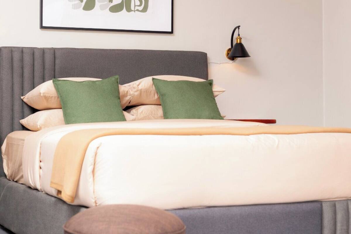 Una cama bien hecha con un cabecero gris, una lámpara de noche y un cuadro en la pared.