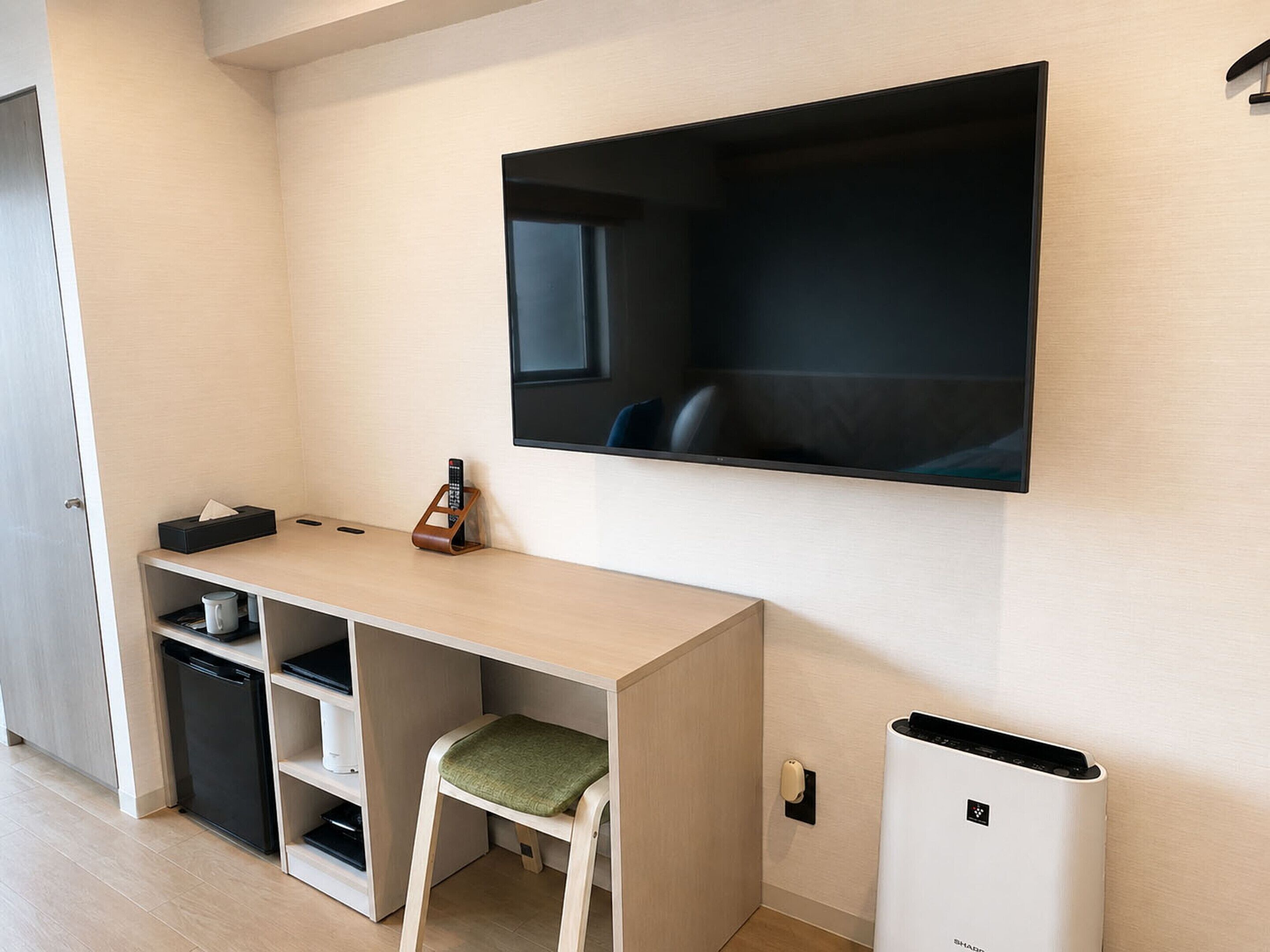 A compact living space with a flat-screen TV mounted on the wall, a small desk with a chair, a trash bin, and a compact refrigerator.