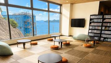 A modern interior with a large window overlooking a body of water and a building, featuring multiple round tables and bean bag chairs.