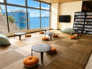 A modern interior with a large window overlooking a body of water and a building, featuring multiple round tables and bean bag chairs. - Livemax Resort Mihara Sea Front (Mihara)