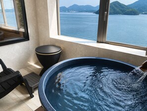 A bathroom with a bathtub, a window with a view of the sea, and a trash bin.