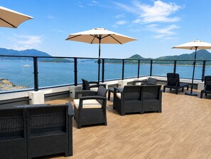 A terrace with wicker furniture and umbrellas overlooking a body of water with mountains in the distance. - Livemax Resort Mihara Sea Front (Mihara)