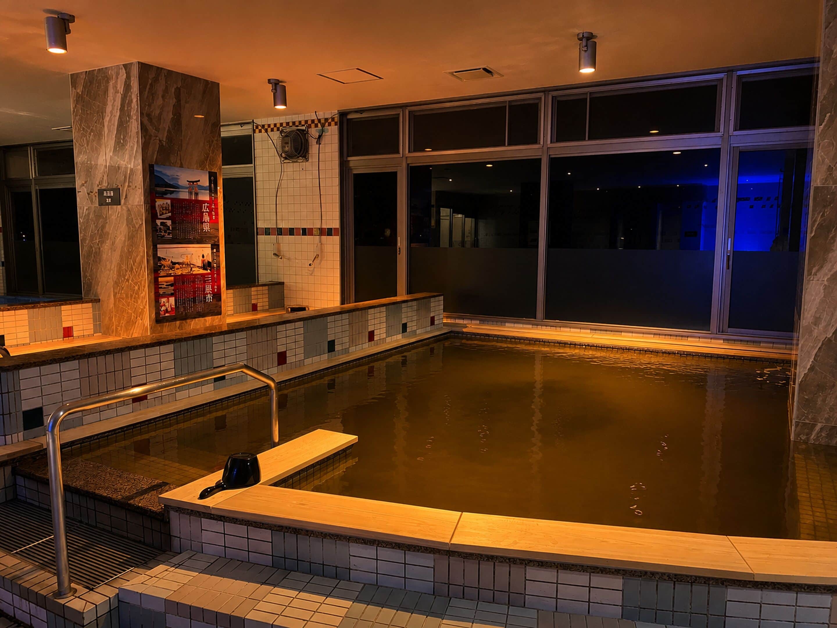 Uma grande piscina interior com borda em azulejo, um pilar de mármore e vista do céu noturno através de amplas janelas.