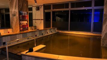 Uma grande piscina interior com borda em azulejo, um pilar de mármore e vista do céu noturno através de amplas janelas.