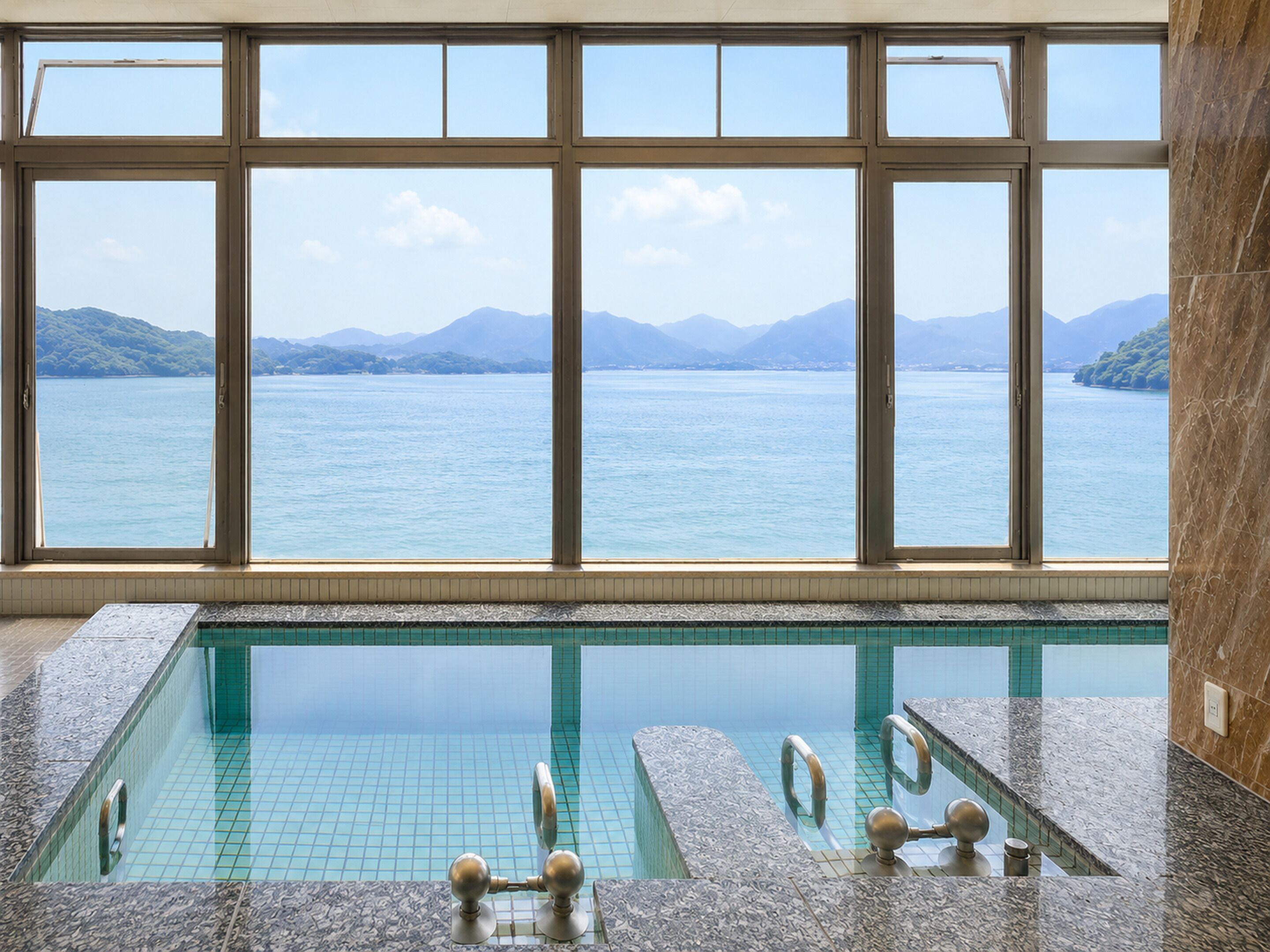 A rectangular indoor pool with clear blue water, surrounded by a tiled floor and walls, with a large window offering a view of a calm lake and distant mountains.