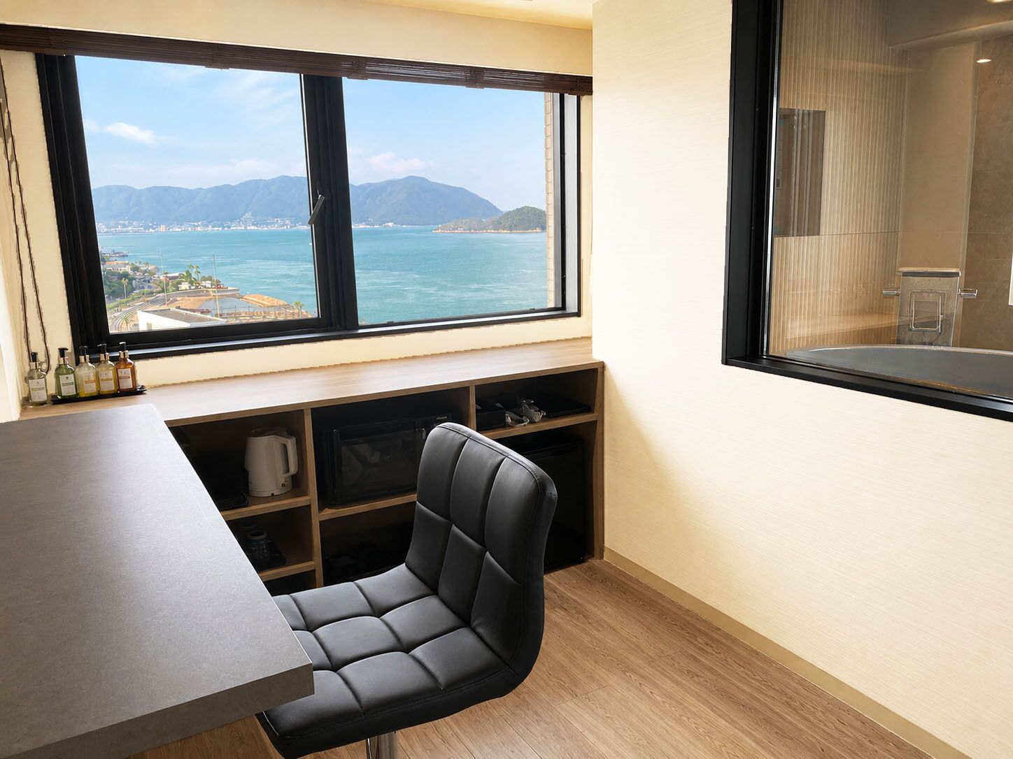Una habitación con un amplio ventanal que ofrece vistas al mar y a las montañas, un escritorio con una silla y un estante con diversos objetos.