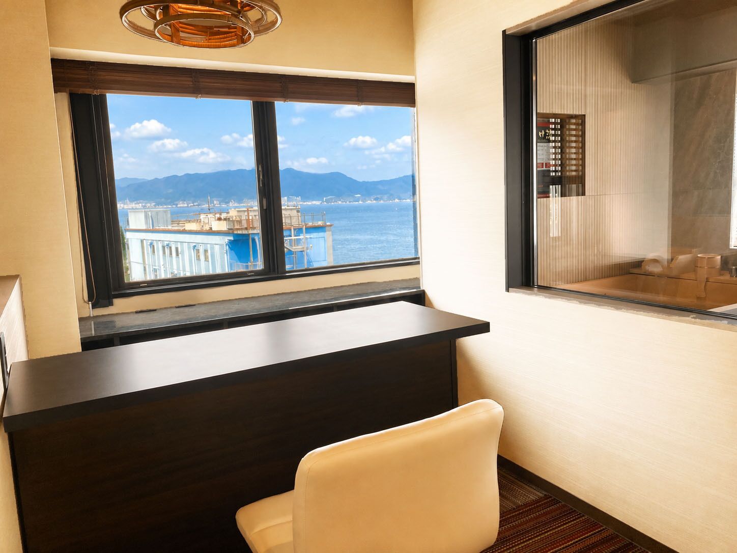 Habitación de hotel con escritorio, una silla, un ventanal con vistas al mar y las montañas, y zona de ducha.