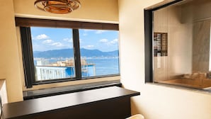 A hotel room with a desk, a chair, a large window with a view of the sea and mountains, and a shower area.