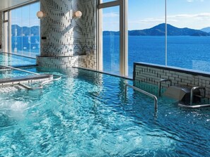 An indoor swimming pool with clear water, a tiled wall, and a view of a body of water and mountains. - Livemax Resort Mihara Sea Front (Mihara)