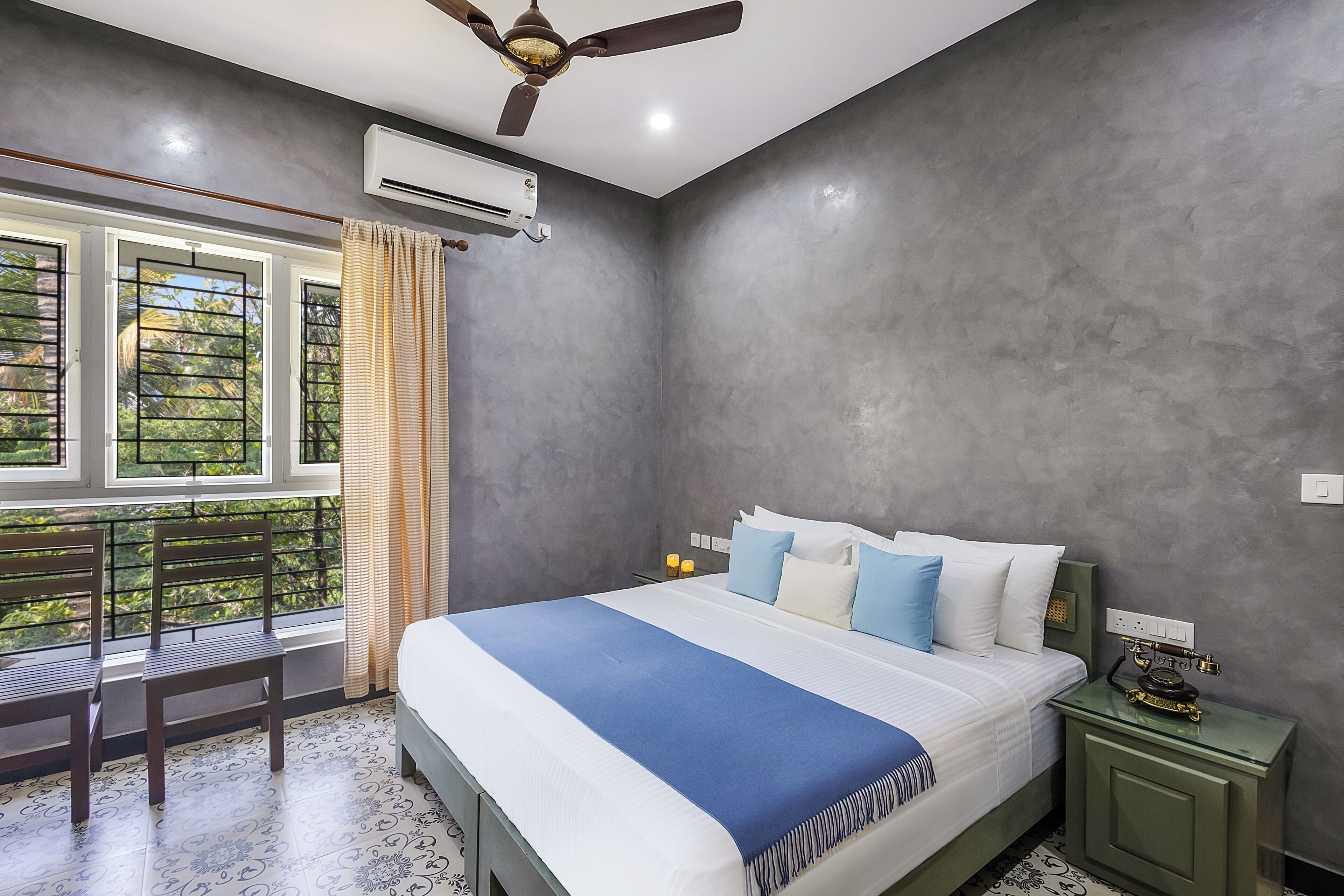 A bedroom with a bed, a nightstand, a ceiling fan, an air conditioner, a window with curtains, and a view of greenery outside.