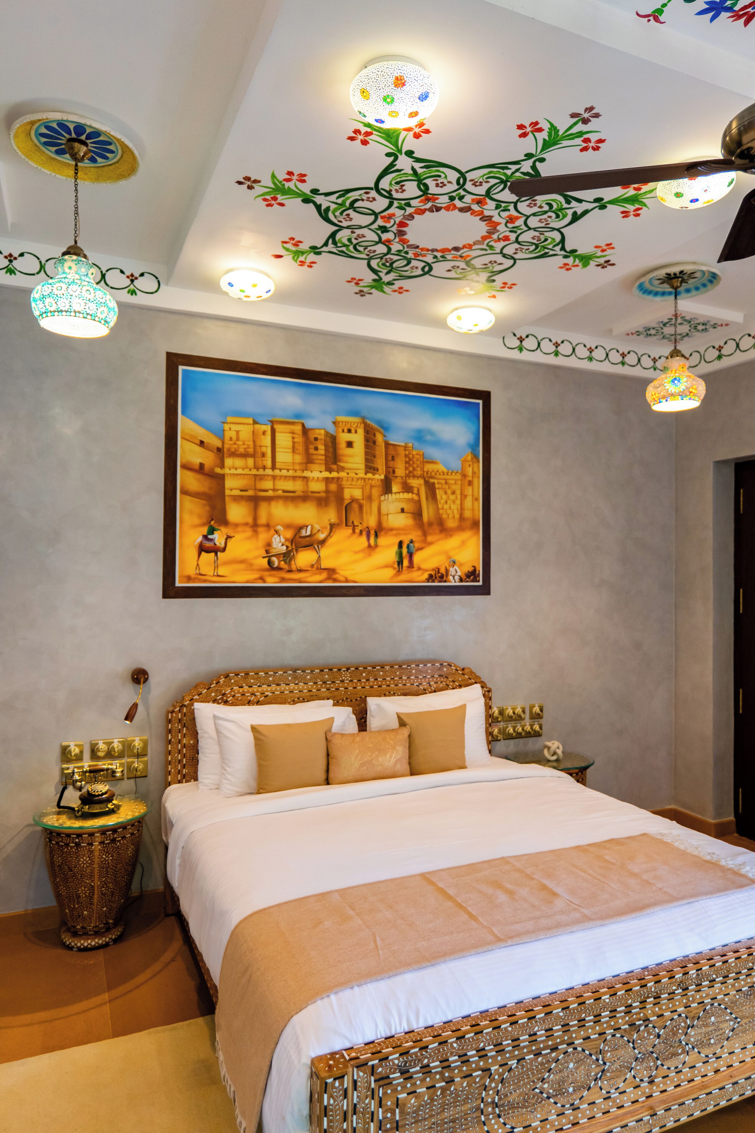 A bedroom with a large bed, a painting of a desert scene, decorative ceiling lights, and a ceiling fan.