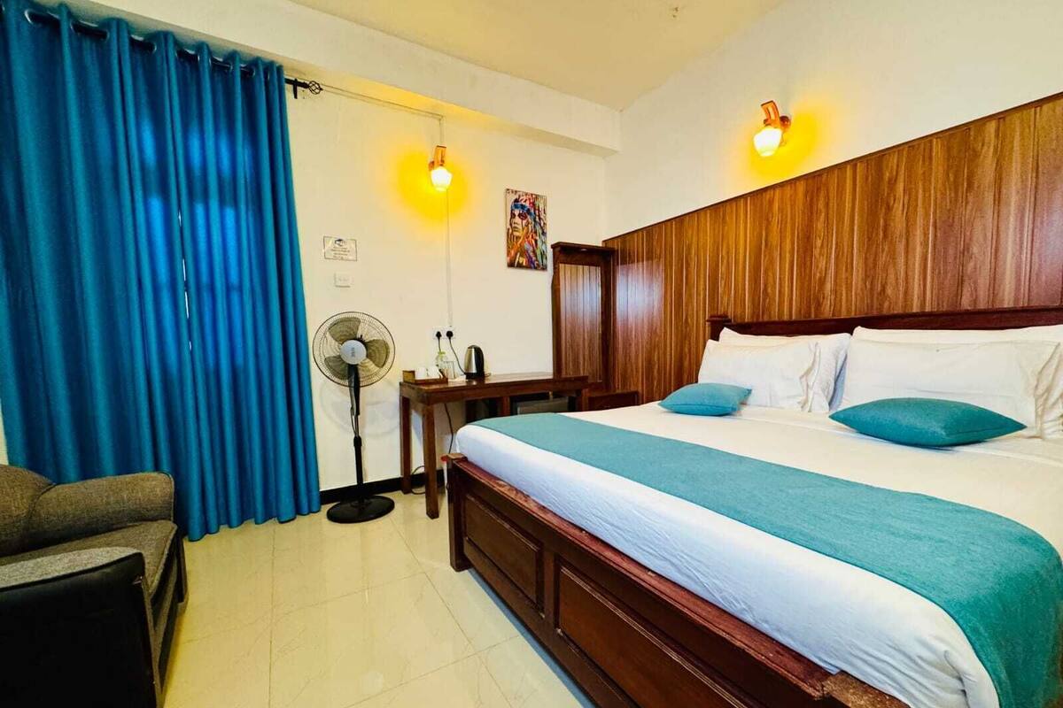 A hotel room with a wooden headboard, a bed with white and teal bedding, a desk with a fan, and a blue curtain.