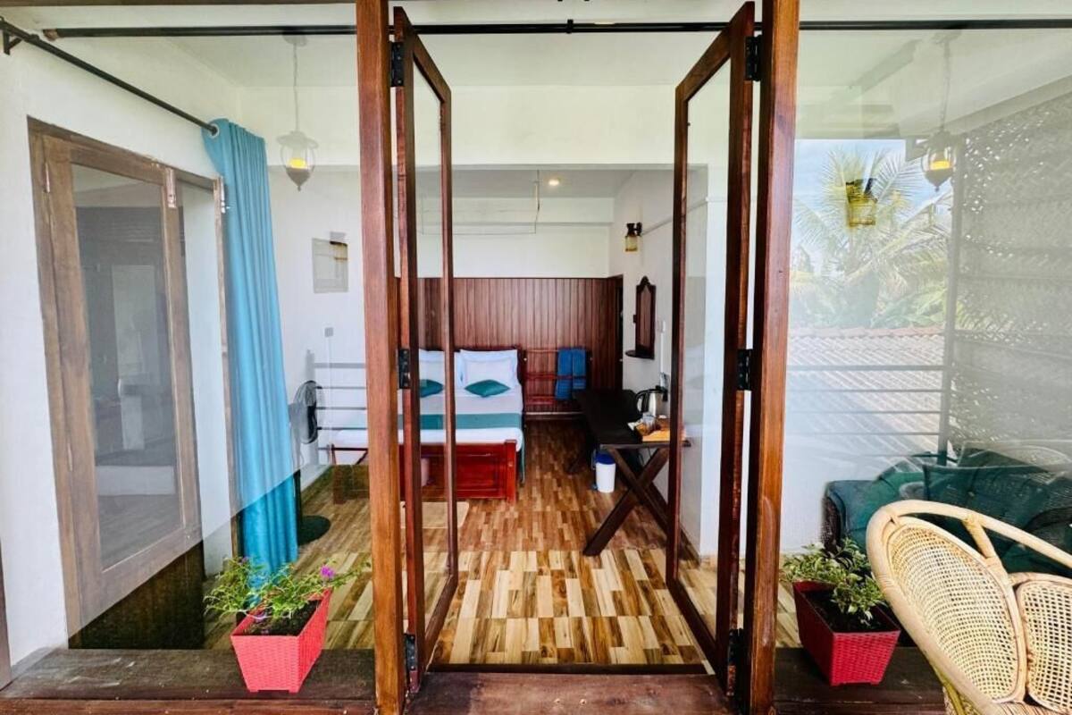 A room with a wooden floor, a bed with a striped pillow, a desk, and a chair. There are two potted plants and a wicker chair.