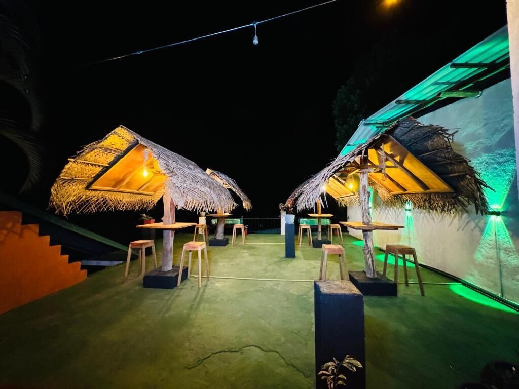 A night scene with two thatched-roof structures, each with a table and chairs, connected by a staircase.