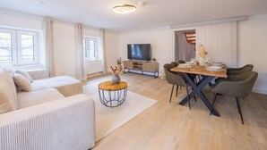 Ein modernes Wohnzimmer mit Sofa, Couchtisch, Esstisch mit Stühlen, Fernseher und einem Fenster mit Vorhängen.