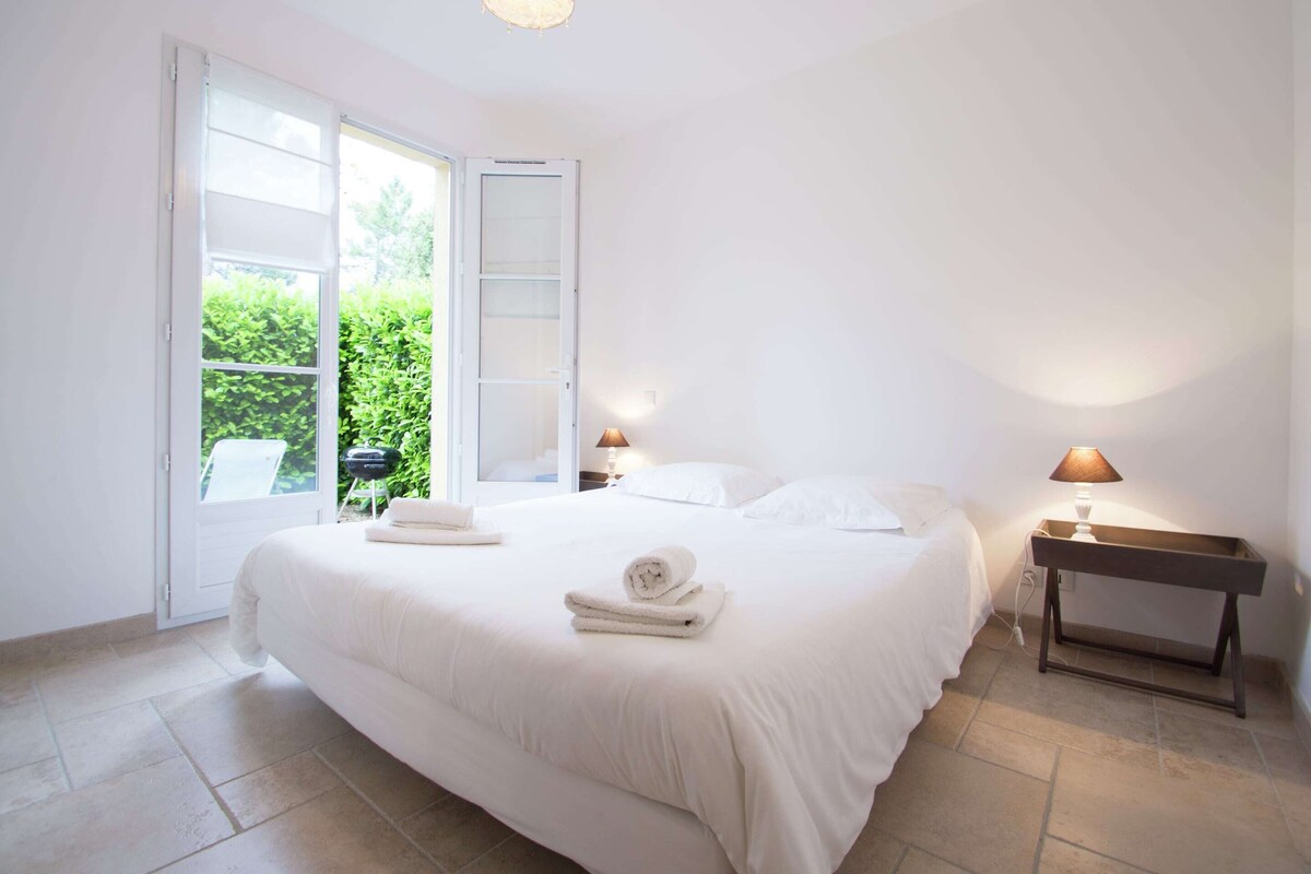 A bedroom with a large bed, two bedside tables with lamps, a sliding glass door to a garden, and a small table with a lamp.