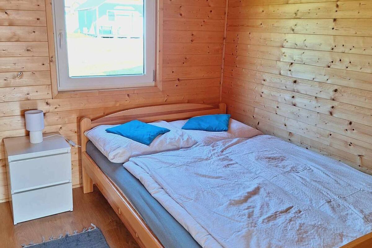 A wooden cabin bedroom with a bed, a nightstand, a lamp, a window, and a painting on the wall.